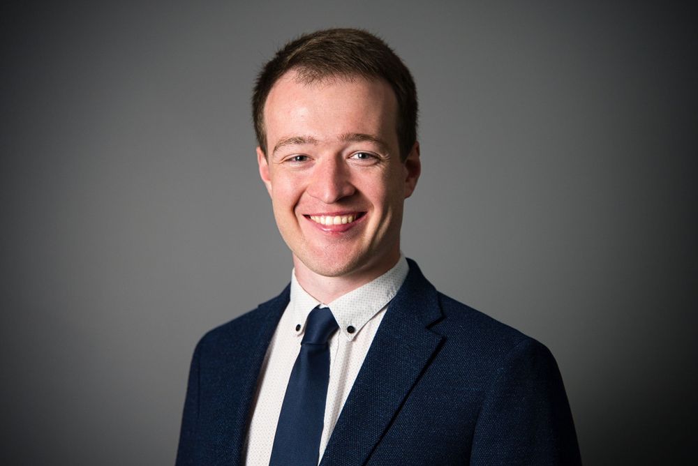 Professional headshot of someone in a navy suit and white shirt against a dark background smiling warmly.