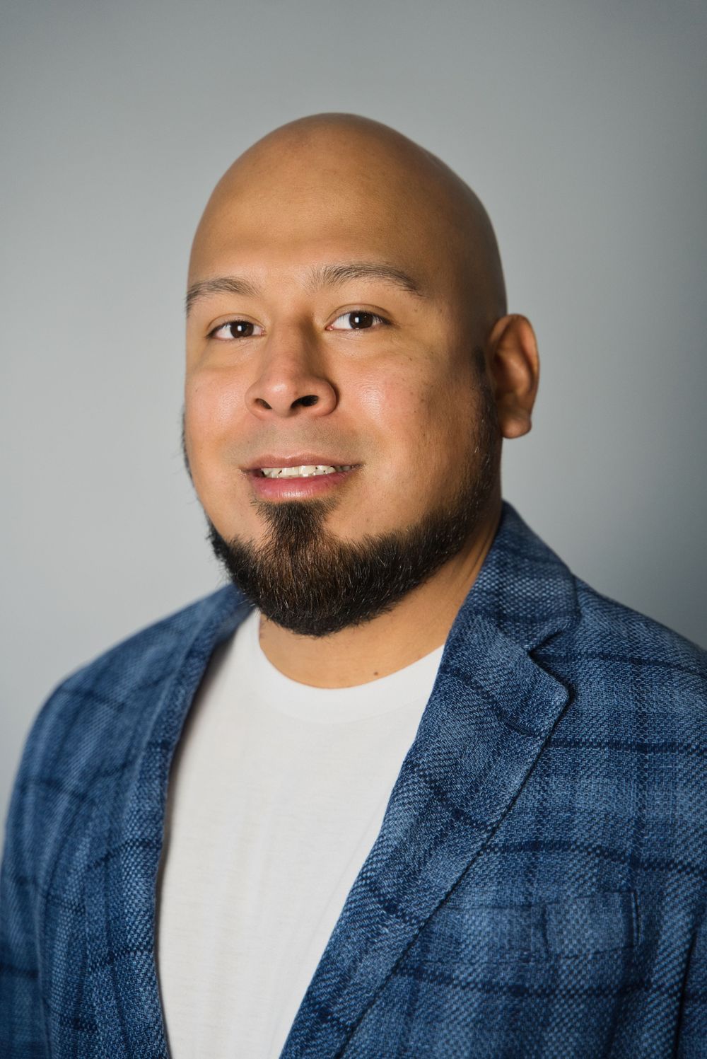 Professional portrait of a person in a plaid blazer with a beard against a grey studio background.