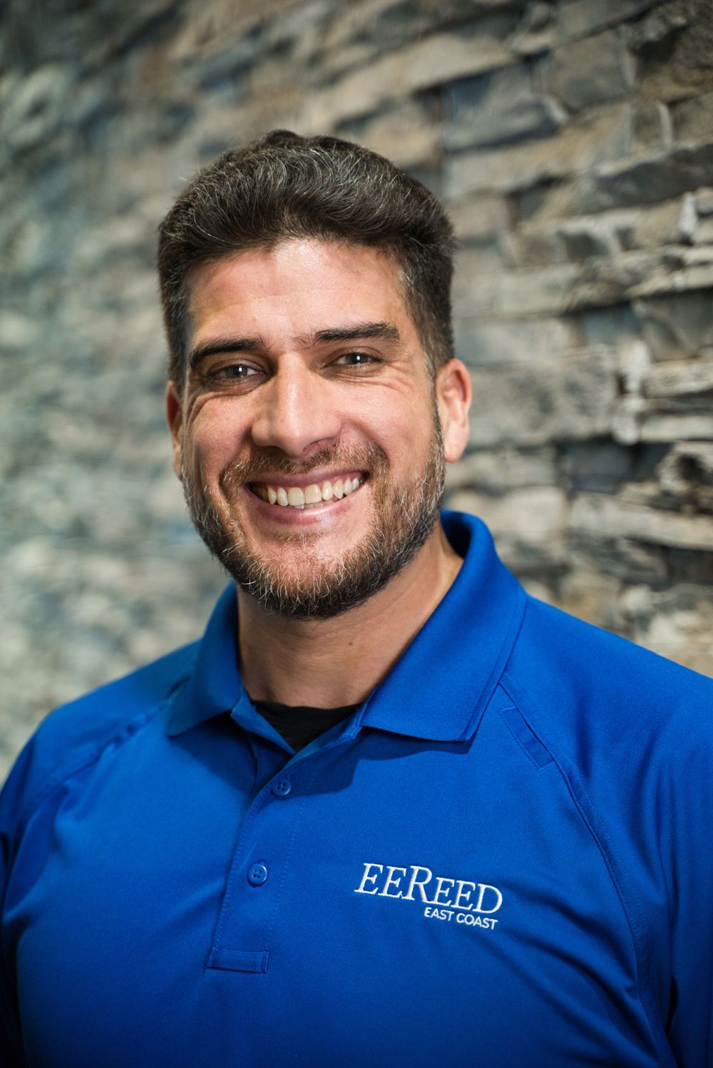 Professional portrait of a person wearing a blue EE Reed polo against a stone wall with a warm smile.