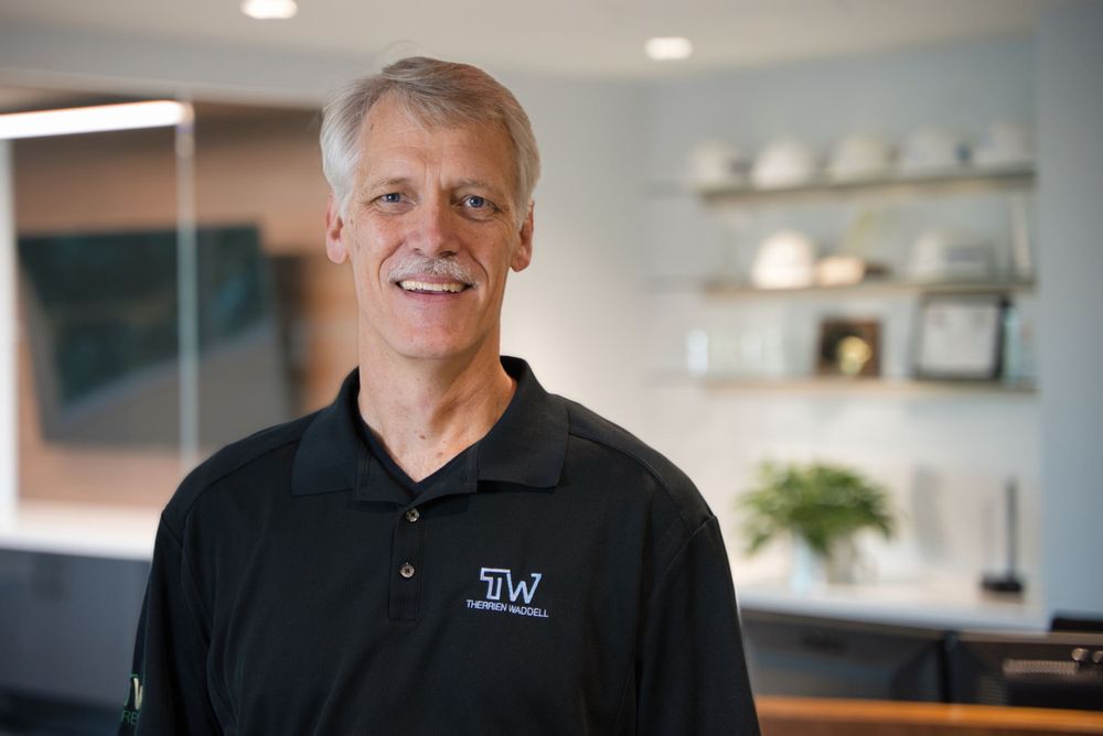 Professional headshot of a person in a black polo in an office setting with awards displayed.