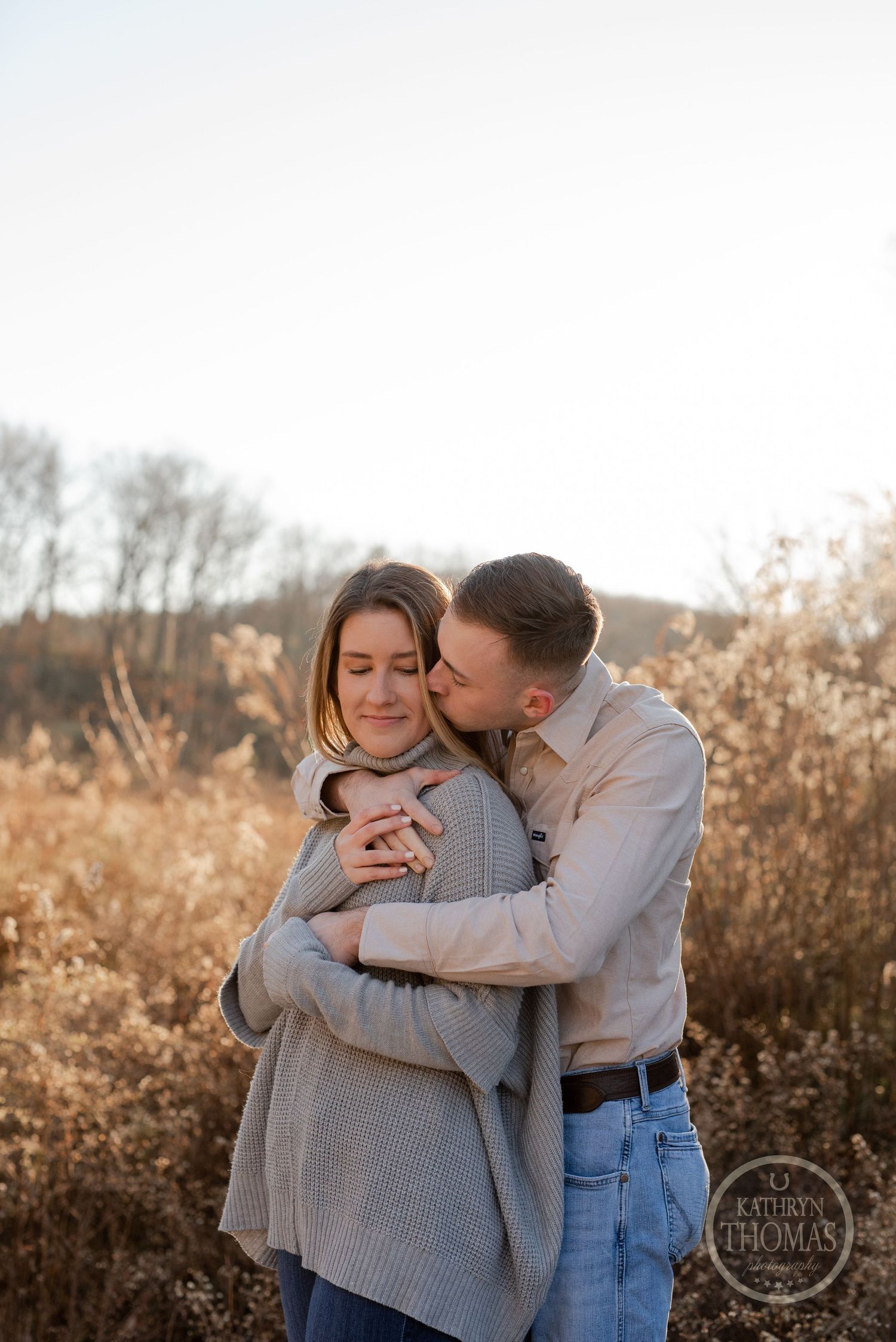 A tender embrace captured in warm sunset light with dried grass and trees creating a natural backdrop.