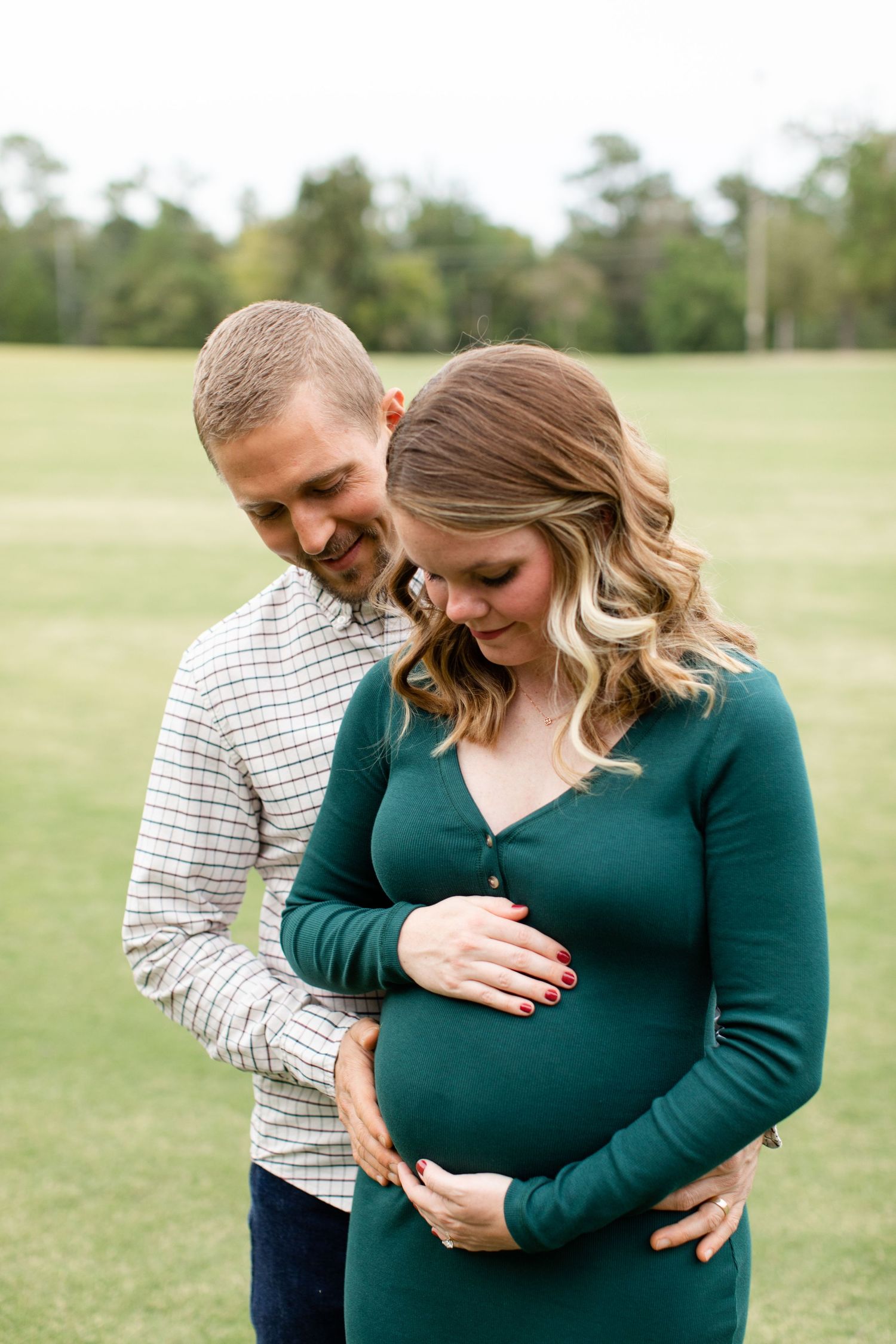 Candid maternity moment in a green field with couple sharing a laugh while holding the baby bump.