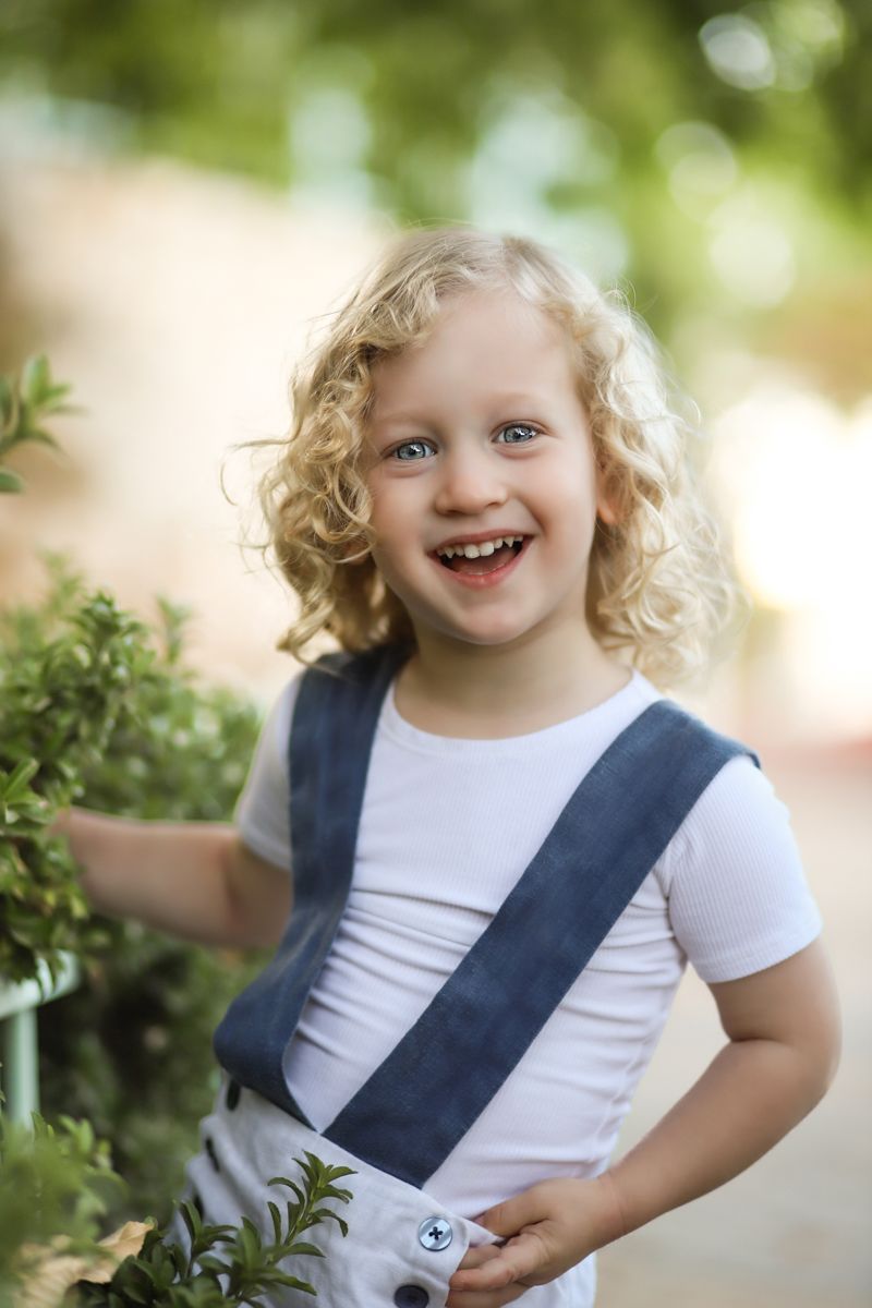 A joyful child with curly blonde hair wearing a white shirt with navy blue overalls stands among green foliage outdoors.