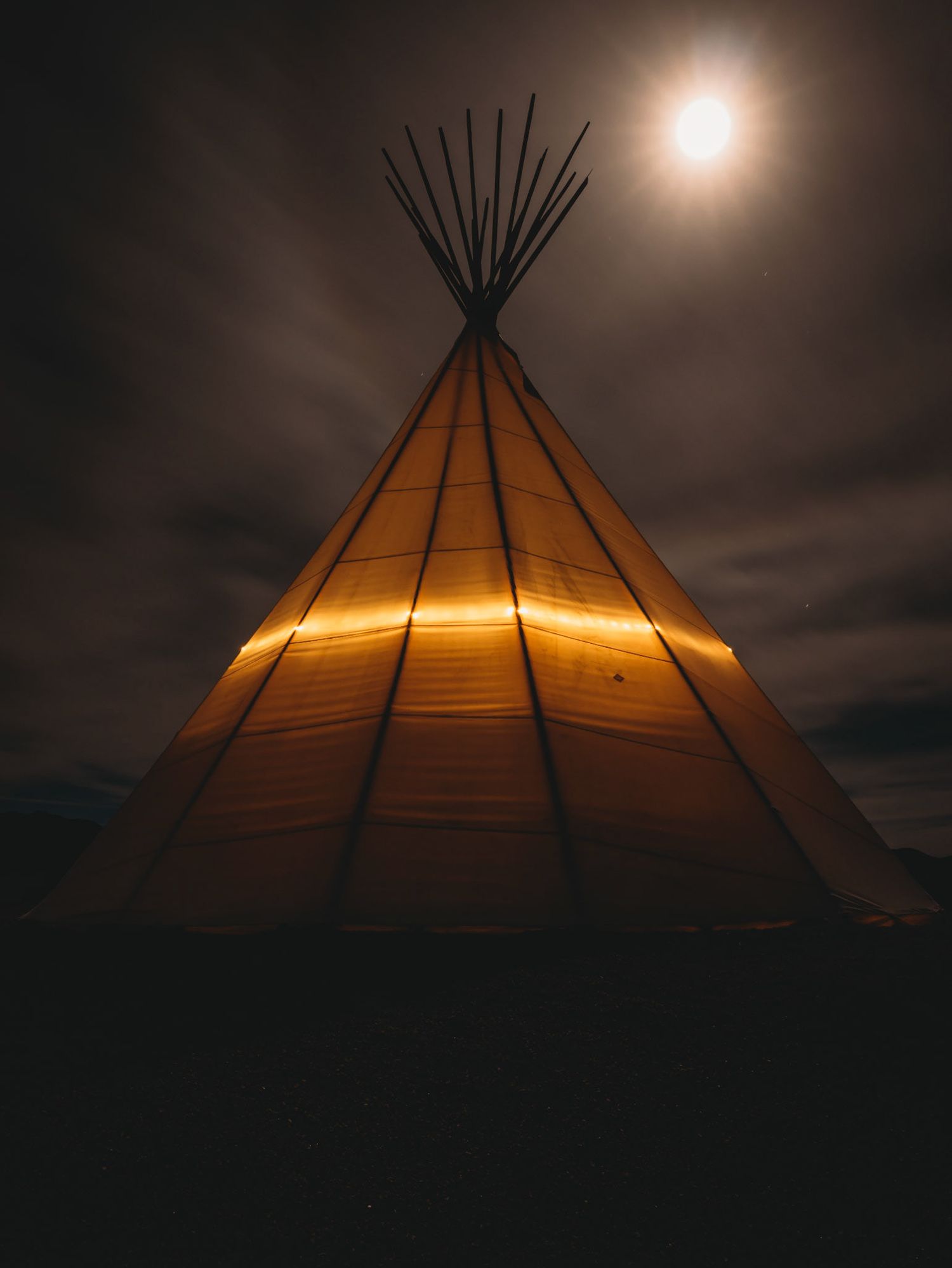 Traditional teepee lit from within against dark night sky with full moon visible.