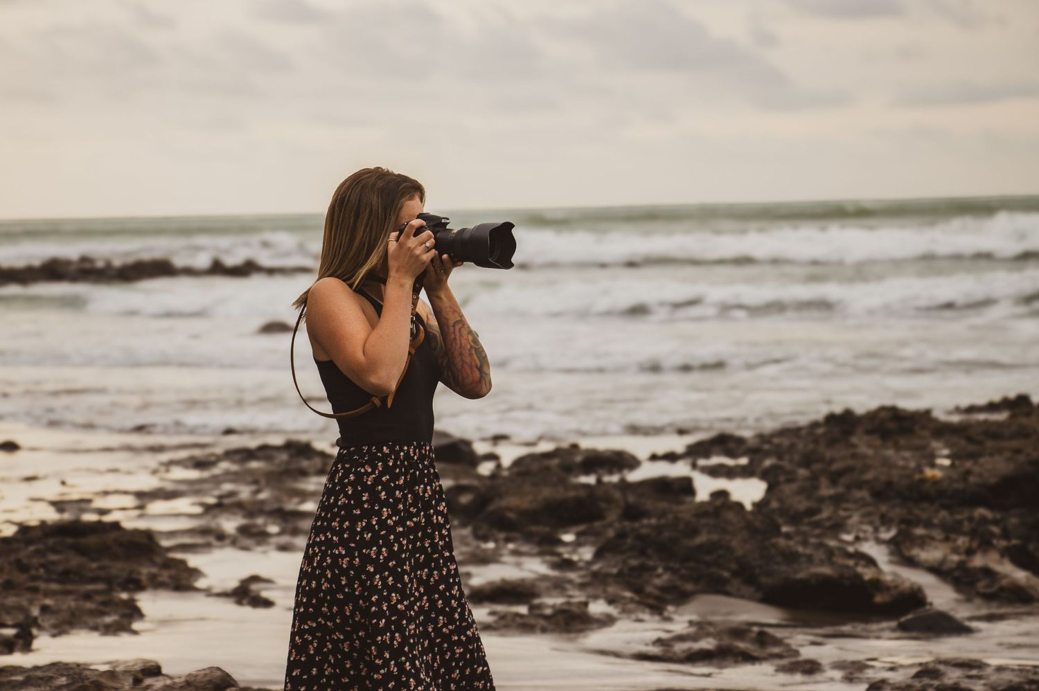 Photographer in floral dress captures ocean waves from rocky shoreline.