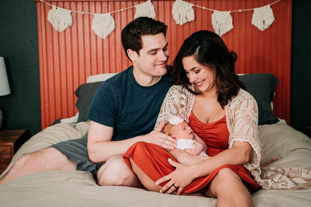 A couple sits together on a bed with paper decorations hanging on the orange wall behind them.