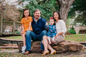 A family of four sits together on a large rock outdoors in a park setting during autumn.