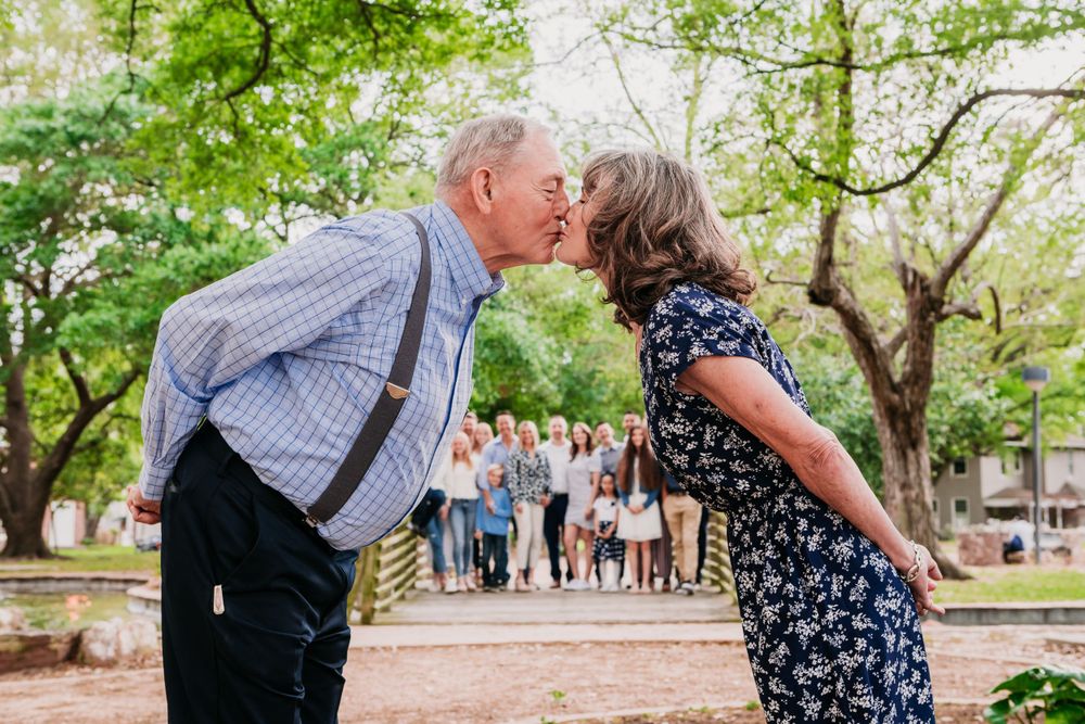 A romantic moment captured between a couple sharing a kiss in a park with a group of onlookers in the background.