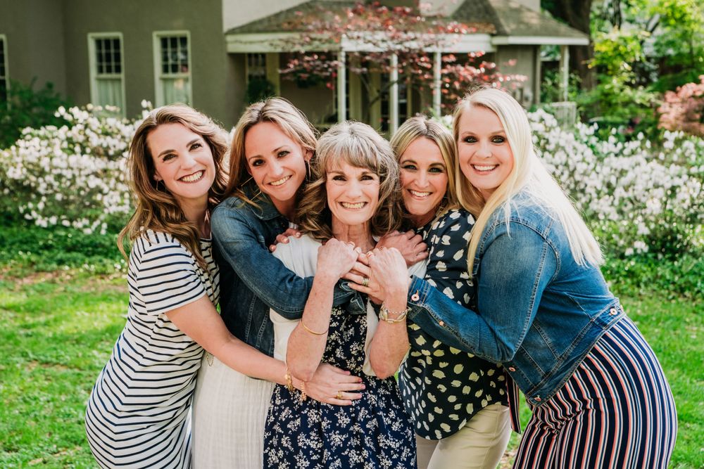 Group in denim jackets and patterned dresses embrace while posing outdoors in front of a garden with white flowers.