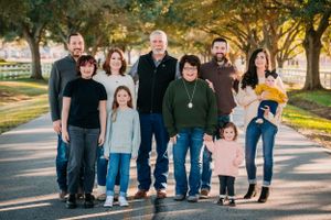 A multi-generational family group stands together on a tree-lined path during a sunny autumn day.