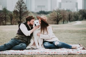 A couple sits on a blanket in a park with their white dog while city buildings rise in the background.