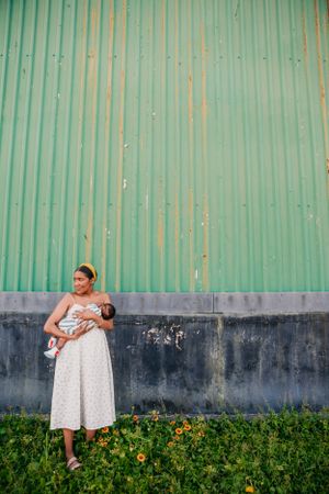 A person in a white polka dot dress stands against a turquoise corrugated wall with yellow flowers growing at the base.