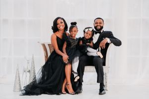 Stylish family wearing elegant black formal attire poses together on white loveseat for studio portrait.