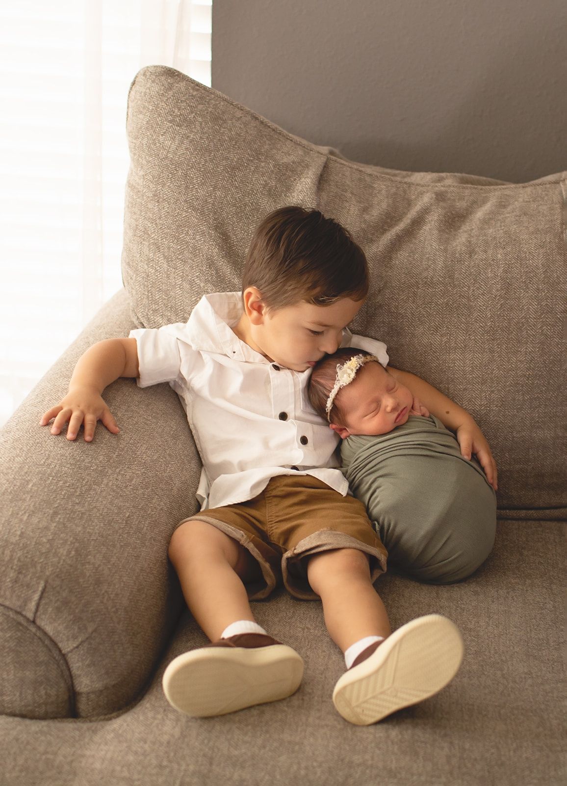 Two children relax together on a cozy gray sofa, with one in a white shirt and tan shorts and the other swaddled in sage green.
