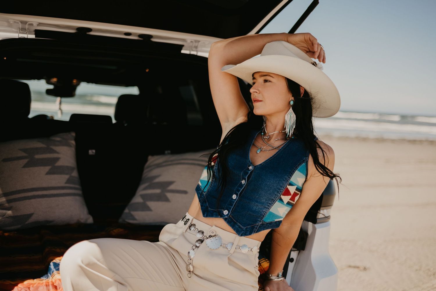 A stylish person in western attire poses against a vehicle at the beach.
