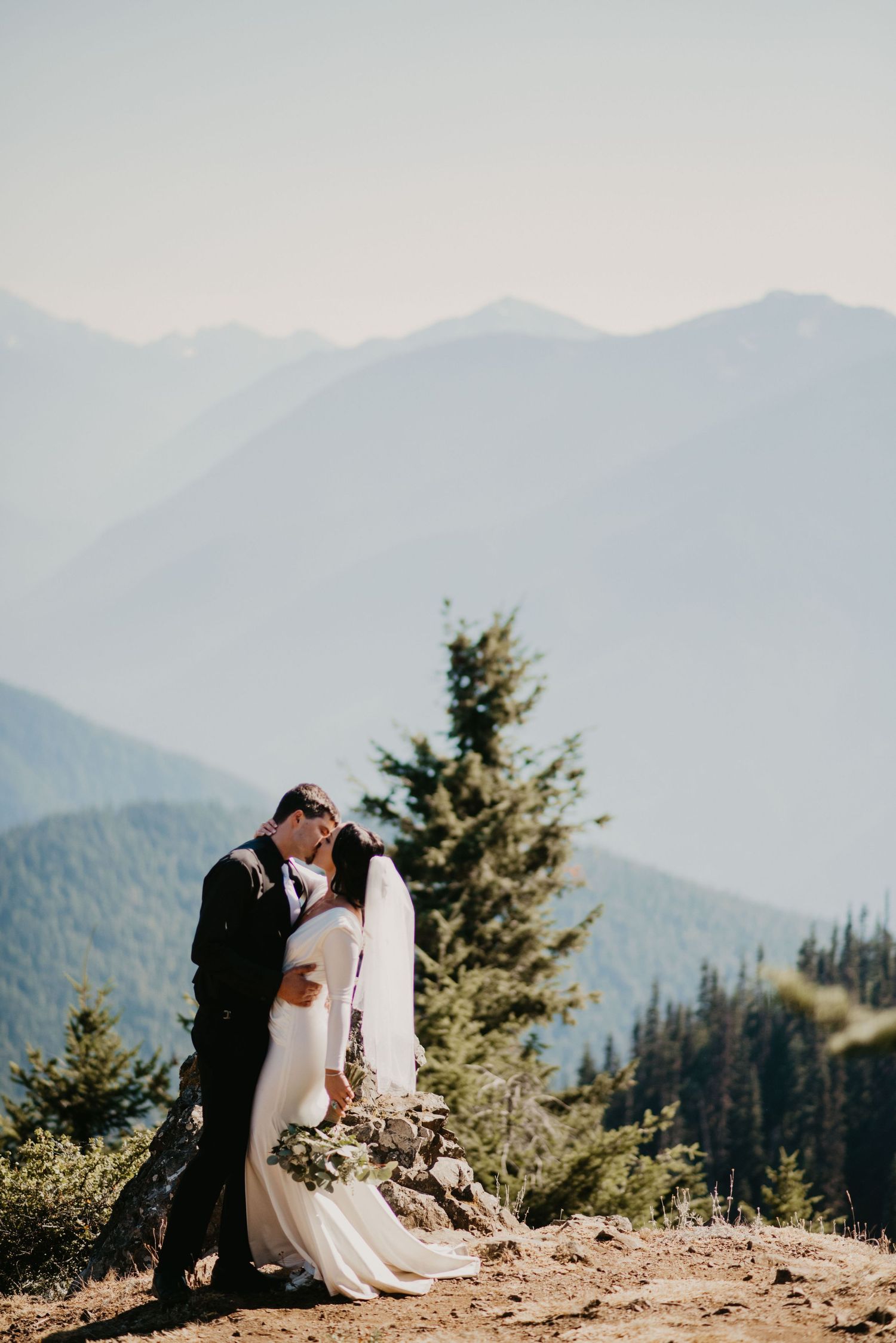 A couple shares an intimate moment on a mountain summit with a dramatic mountain range backdrop.