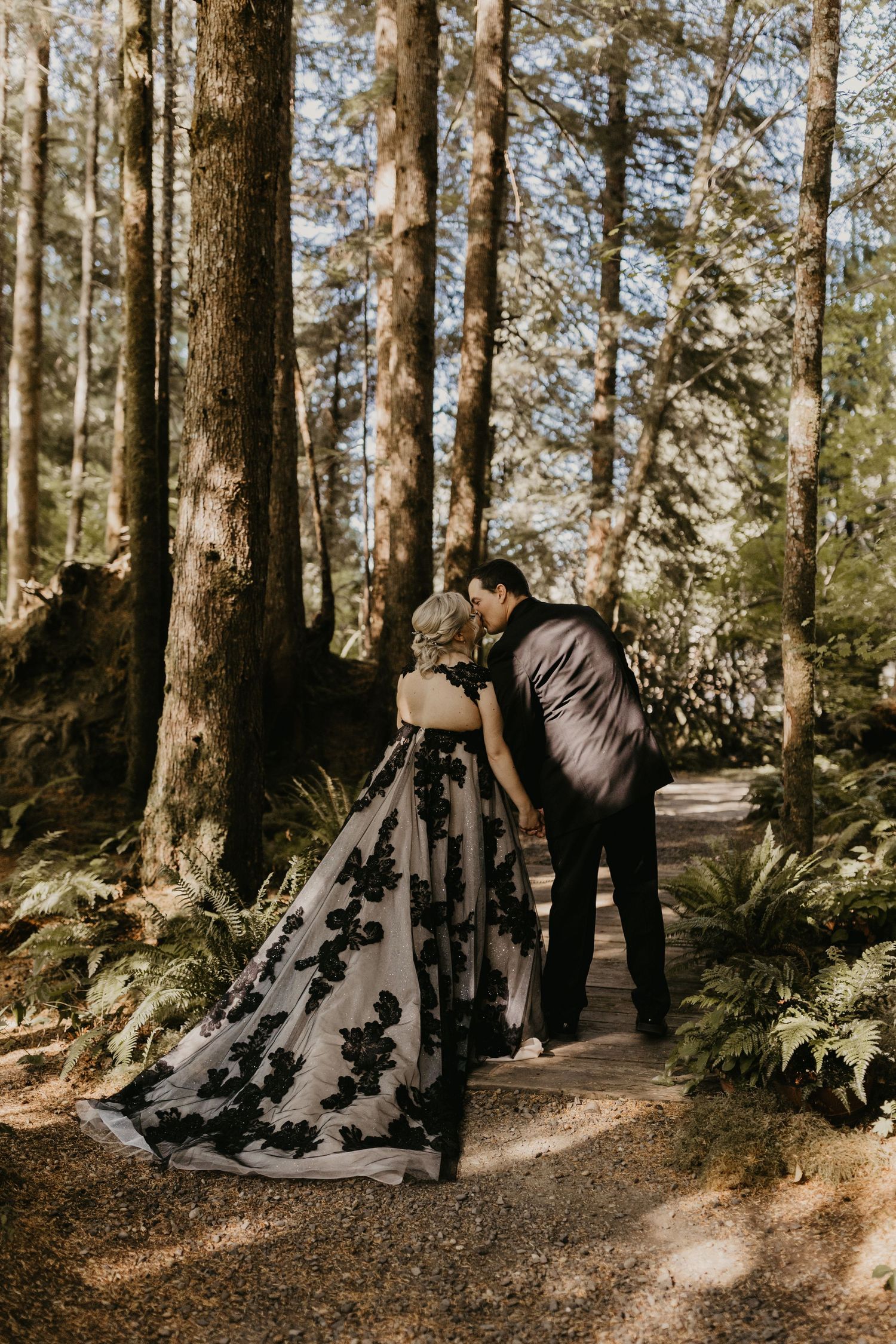Couple in formal wear sharing an intimate moment on a forest path surrounded by tall trees.