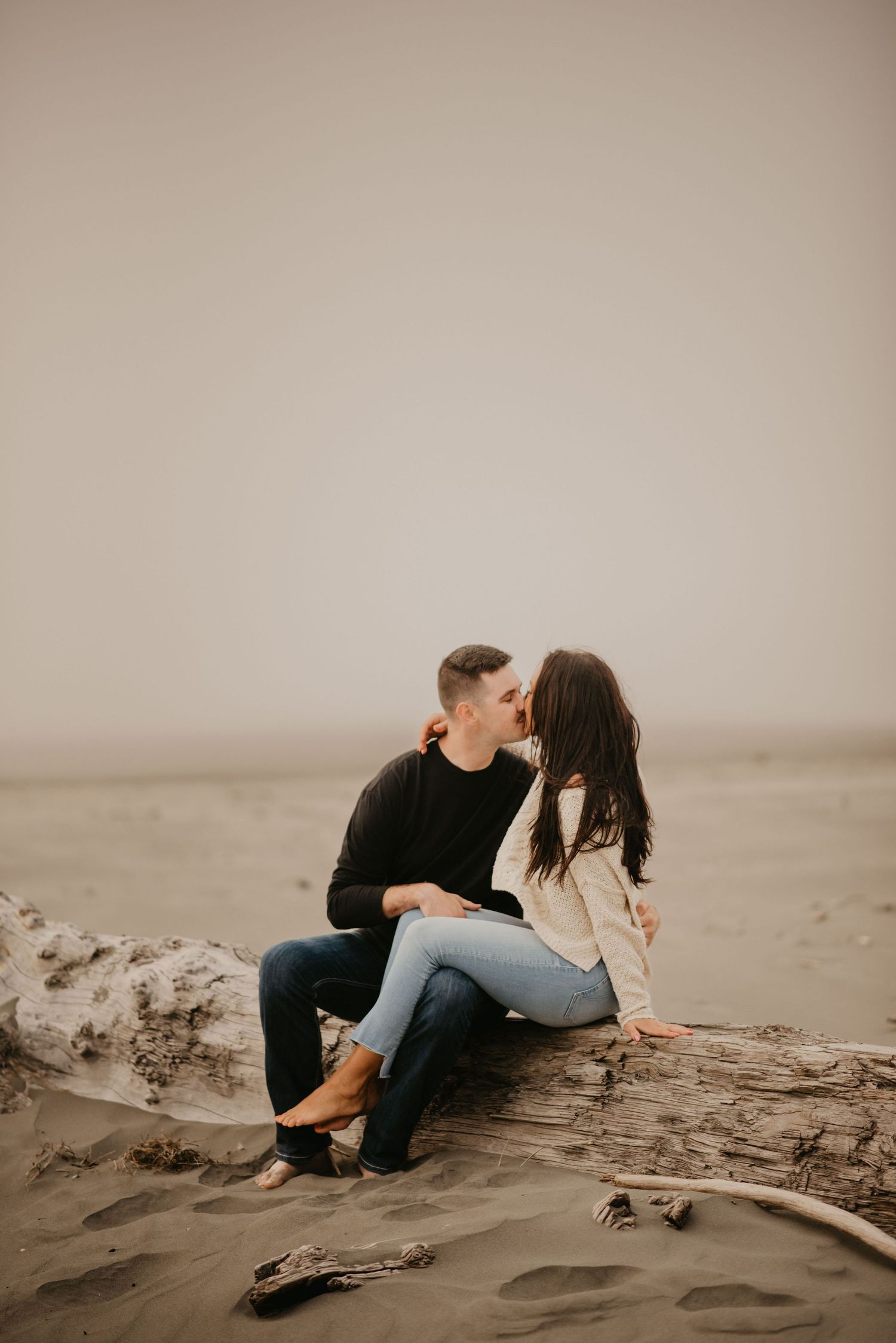 A couple shares a tender moment while sitting on driftwood at a misty beach.