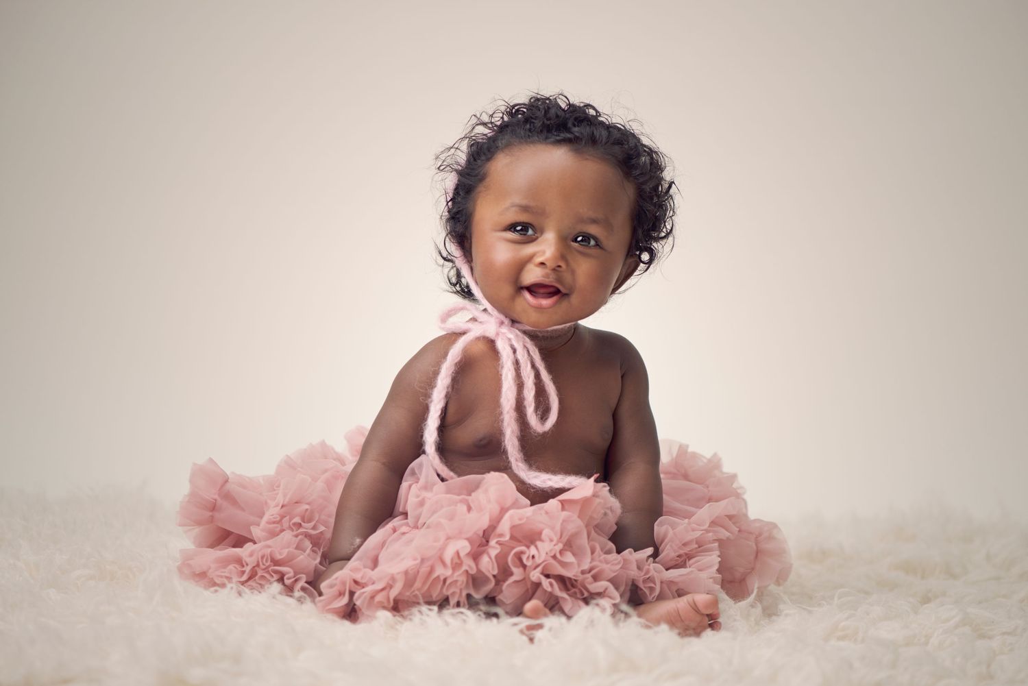 A happy baby wearing a pink tutu sits on white fur during a studio portrait session.