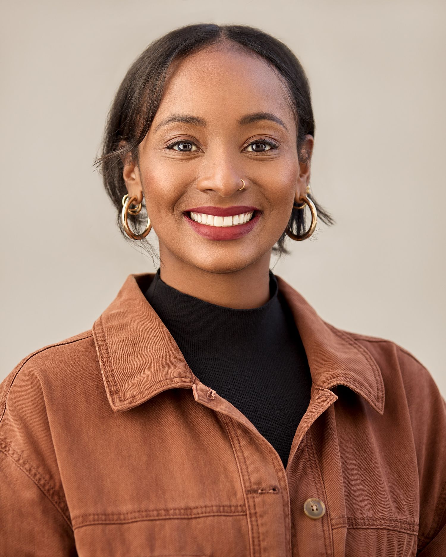Portrait of person in brown jacket and black turtleneck with gold hoop earrings.