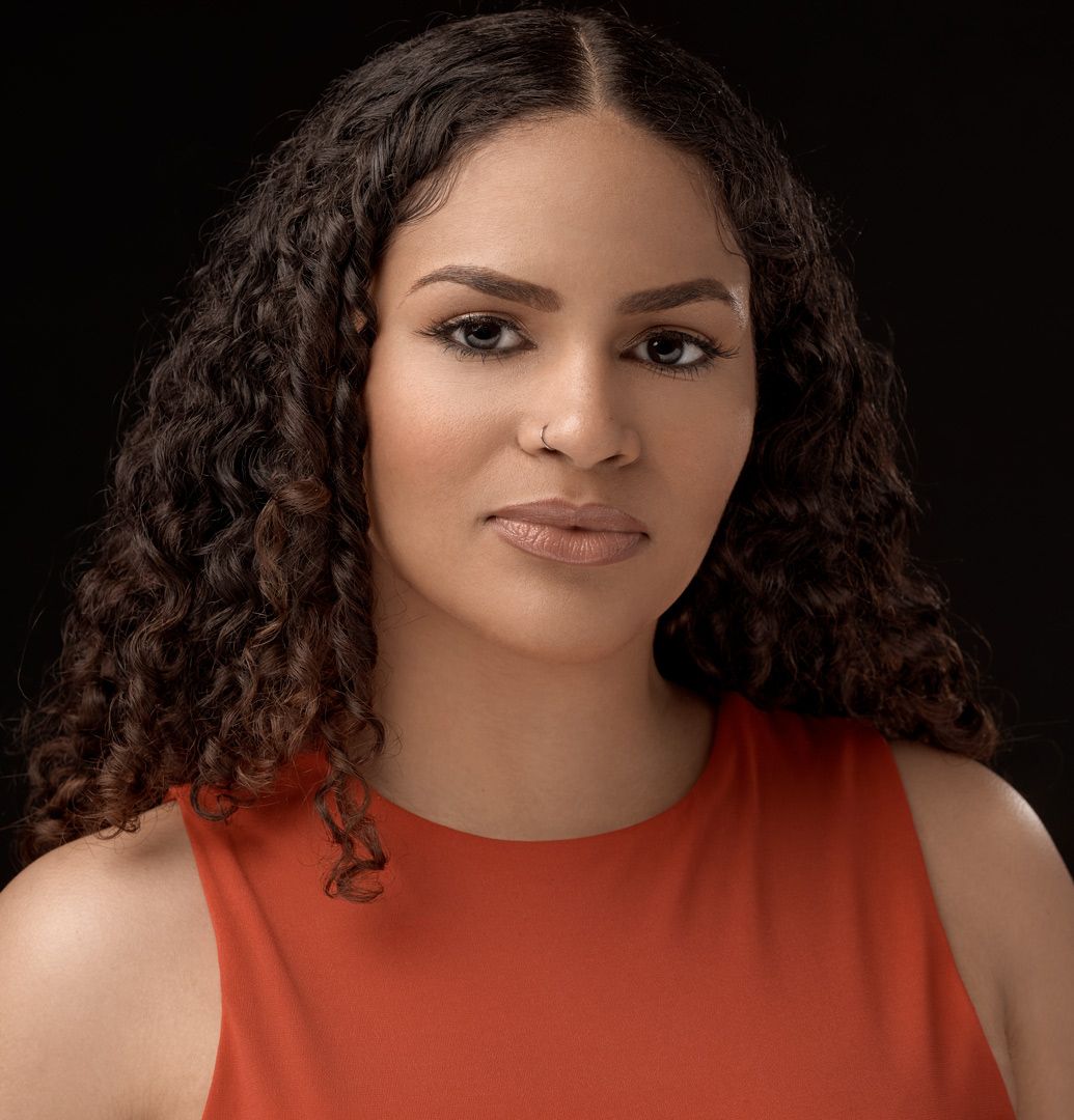 Professional studio portrait featuring glowing skin and natural curly hair against dark background with orange top.