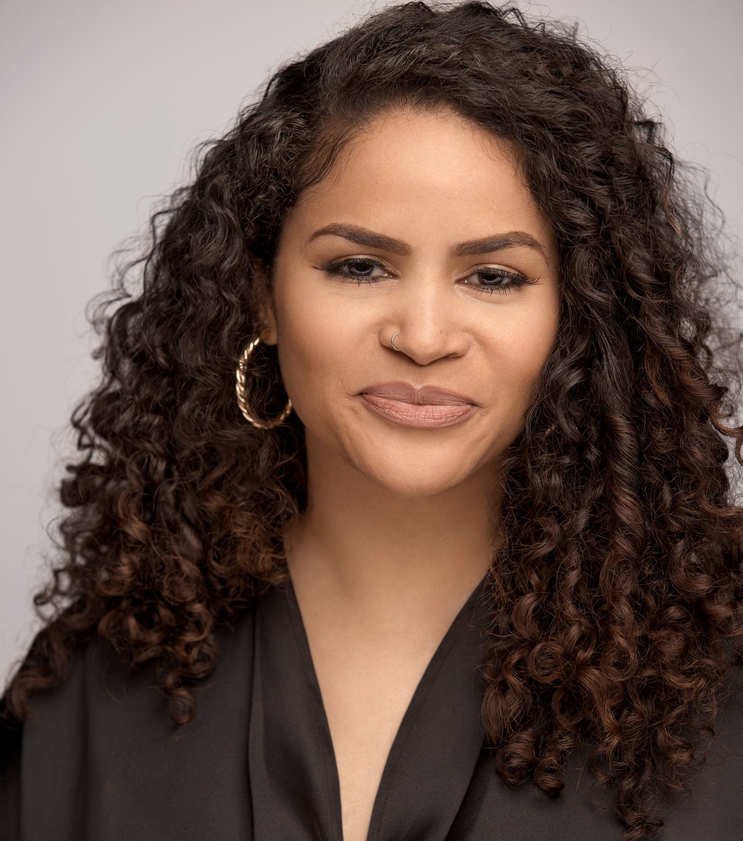 Elegant headshot with long curly hair wearing black top and gold hoop earrings against light backdrop.