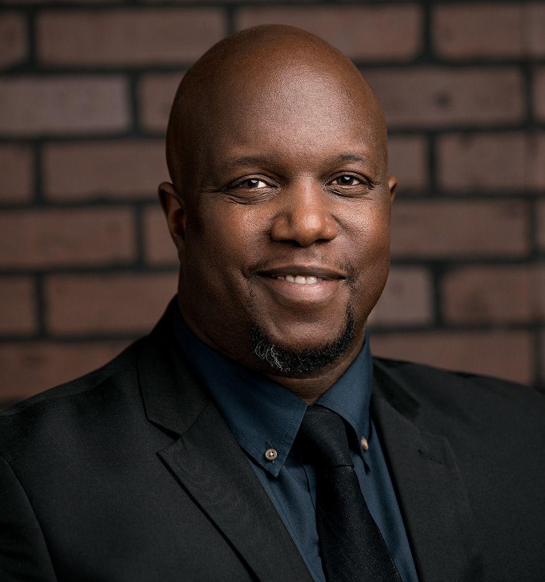 Professional headshot of person in dark suit and dress shirt against brick wall background.