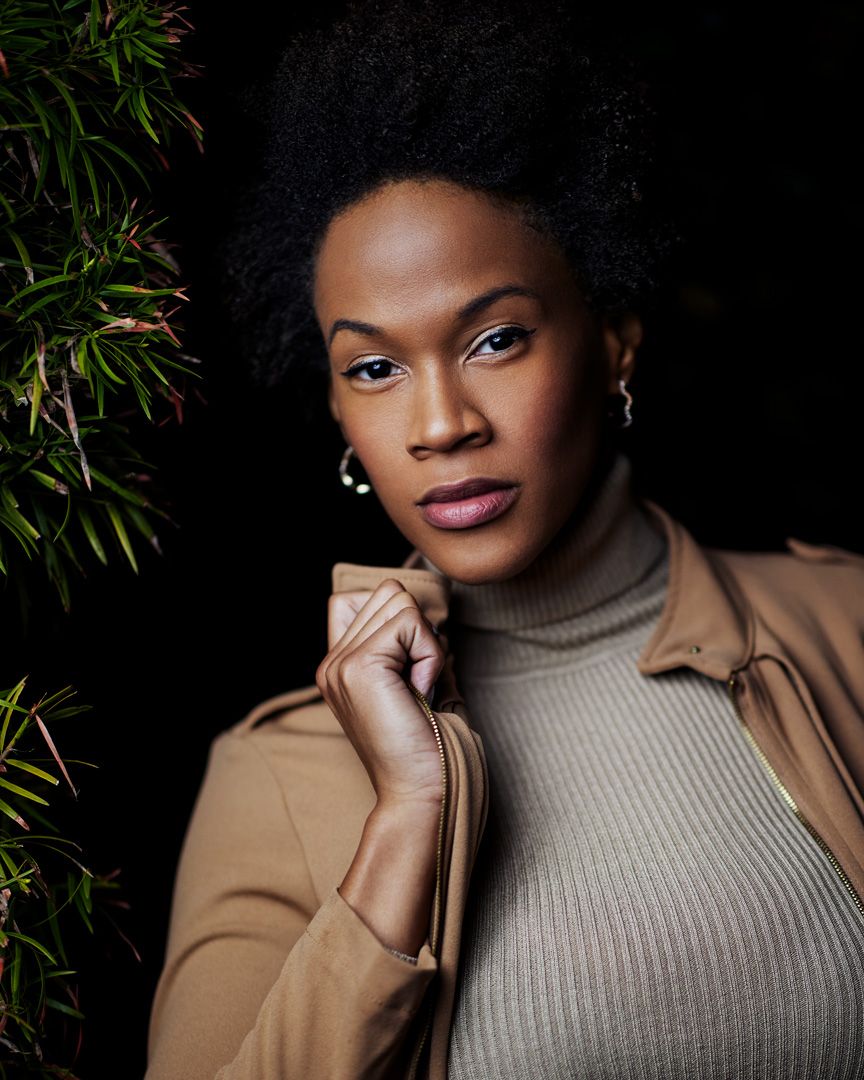 Professional portrait of a person in turtleneck and tan coat against dark background.