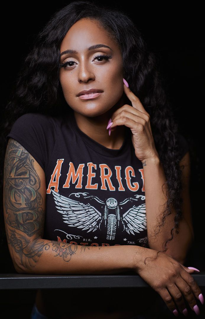 Studio portrait of a person wearing an America graphic t-shirt with visible arm tattoos.