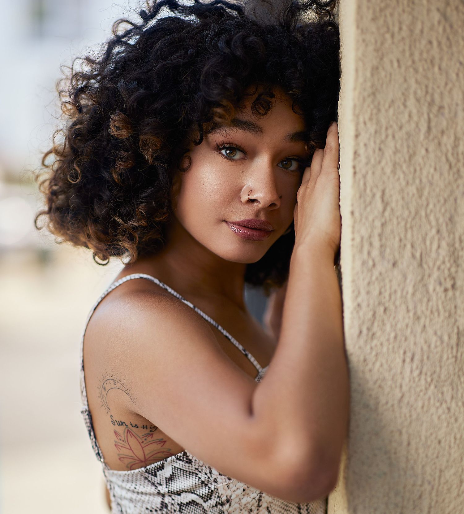 Professional portrait of a woman with natural curly hair wearing a patterned top against a light wall.