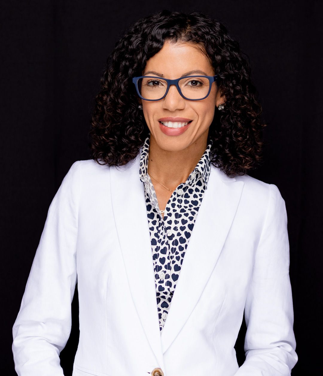 Professional portrait in white blazer and patterned blouse against dark background.