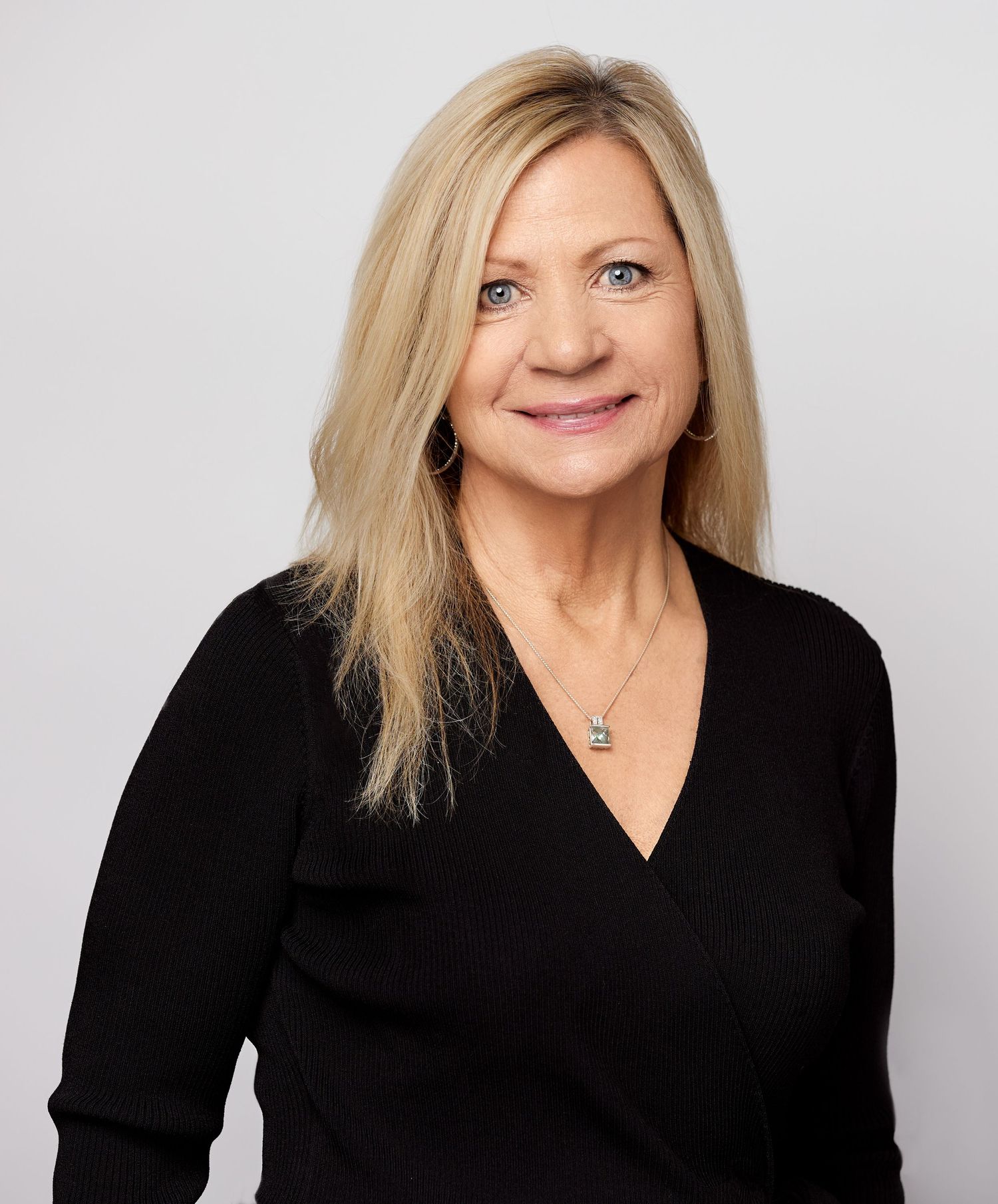 Professional portrait of a person in black v-neck top with blonde hair against grey background.