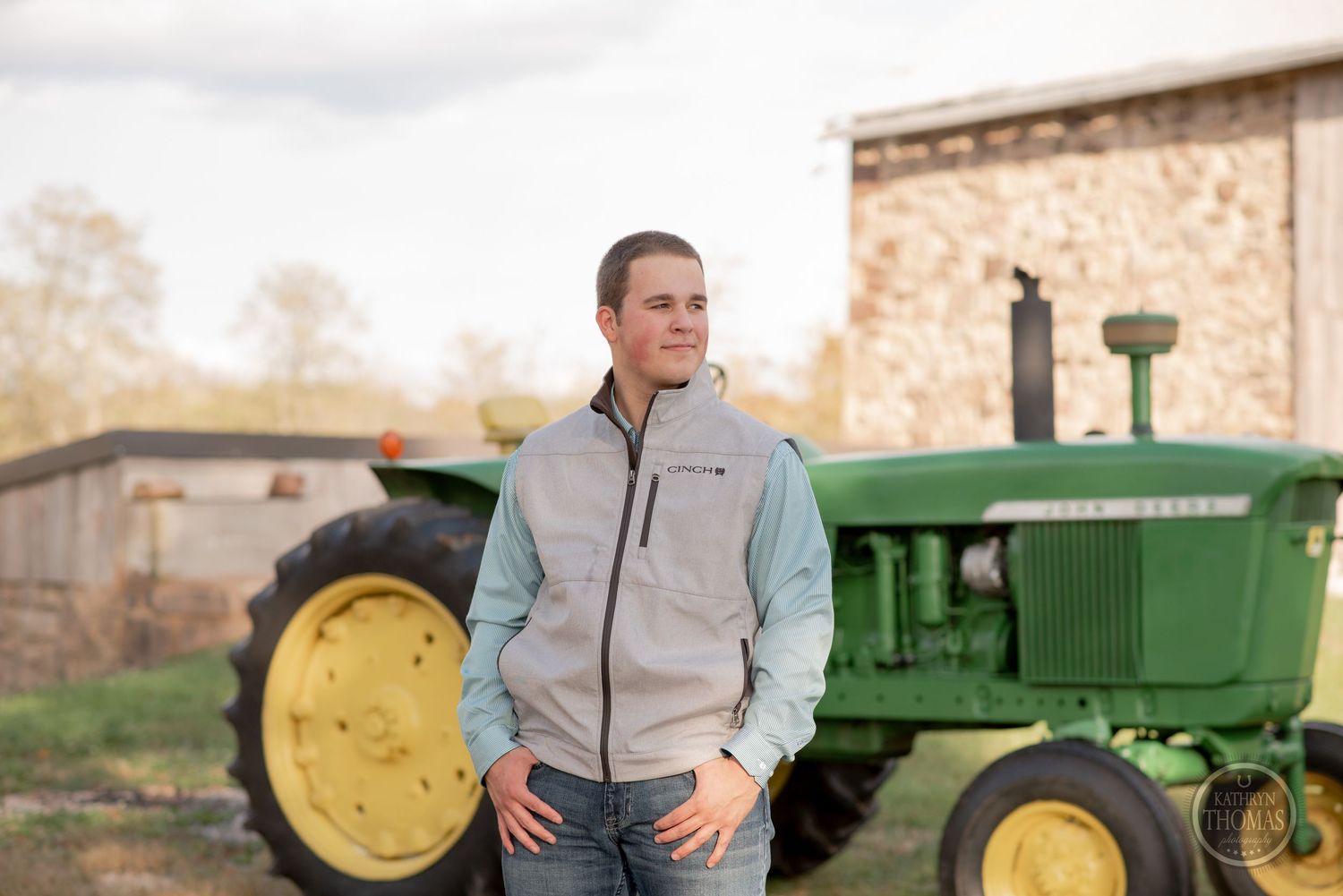 Person in casual wear stands confidently next to vintage green John Deere tractor by stone barn.