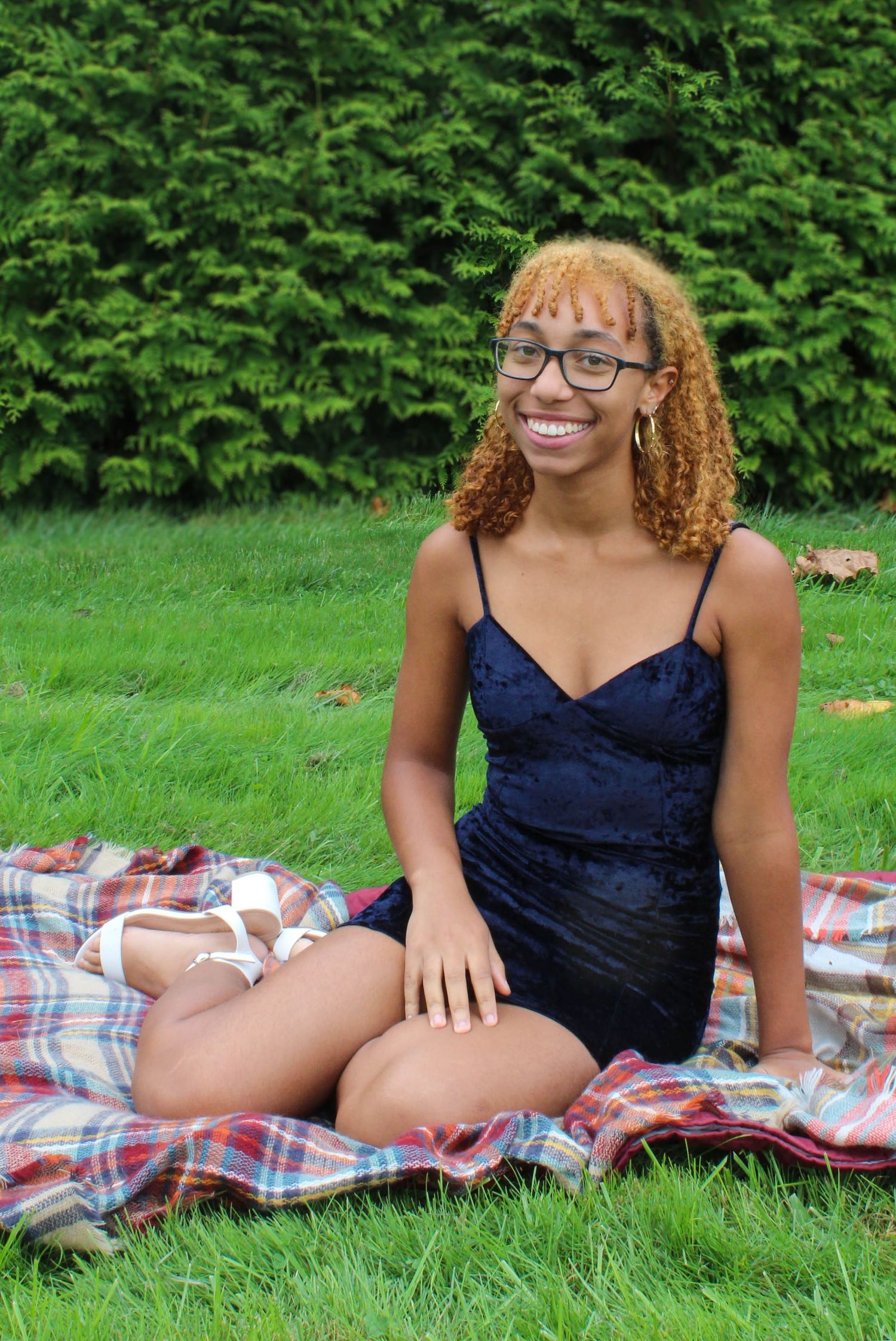 Person in a navy dress sitting on plaid blanket against green hedge background.