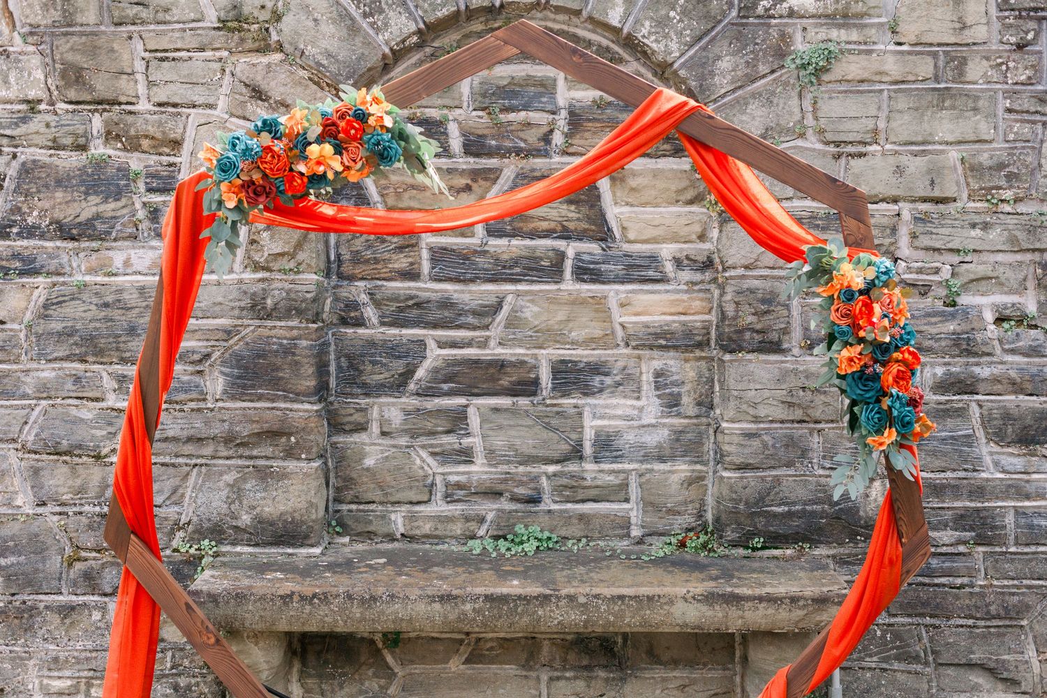 Stone wall decorated with orange flowers and red ribbons for a rustic wedding ceremony backdrop.