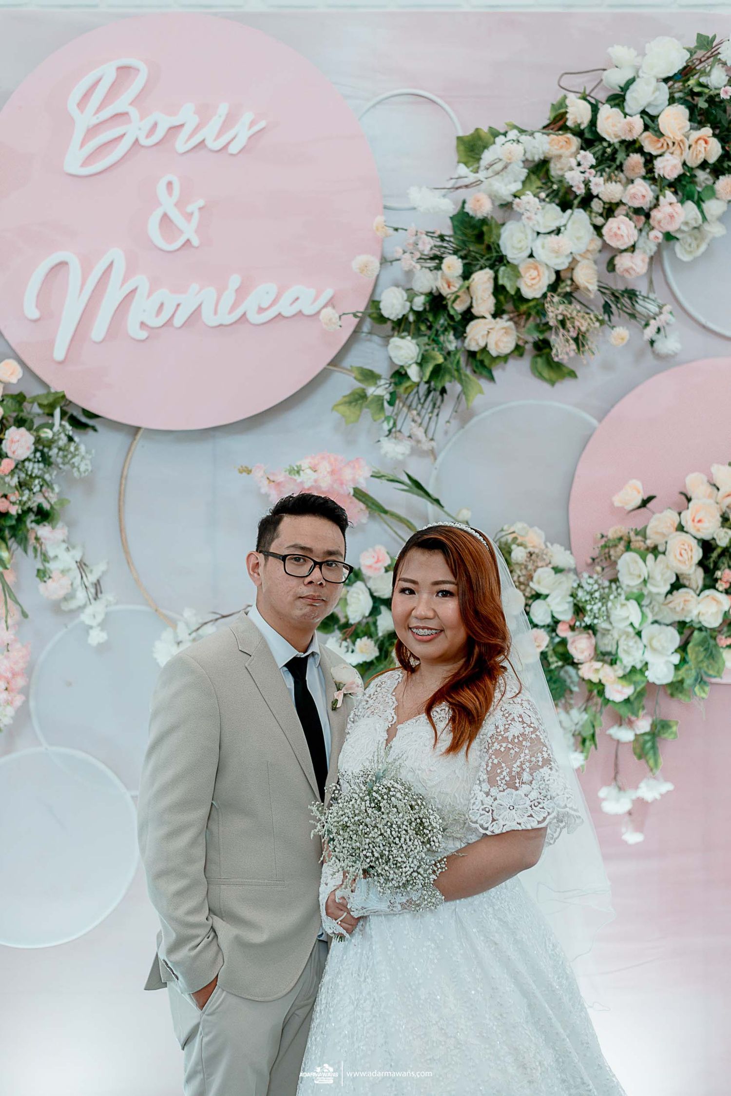 Romantic wedding portrait against pink circular backdrop with Bess & Monica signage and floral decorations.