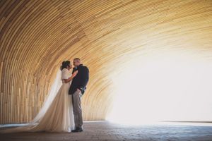 Bride and groom sharing romantic embrace inside stunning curved wooden tunnel architecture.