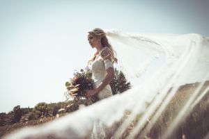 Bride in white gown with flowing veil holding bouquet outdoors on sunny day.