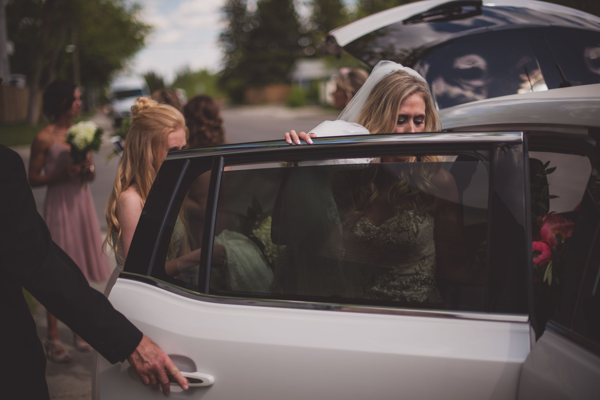 Bride in white dress getting into white limousine surrounded by wedding guests.