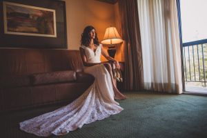 Bride in lace wedding gown sitting on hotel sofa by window with warm lamp light.