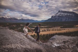 Bride and groom hold hands while climbing rocky mountain cliff with dramatic valley landscape behind them.