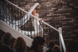 Elegant bride in fitted lace gown descending staircase with guests watching below at wedding venue.