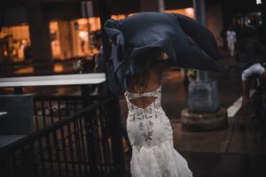 Bride in lace wedding gown shielded by large black umbrella outside rainy wedding venue.