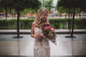 Blonde bride in floral wedding gown holding vibrant pink bouquet outdoors at urban venue.