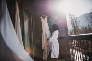 Bride in white robe admiring wedding gown hanging on sunlit mountain chalet balcony.