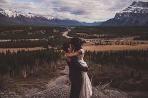 Newlyweds share a loving embrace on a rocky overlook with a winding river valley below.