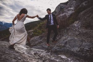 Groom helps bride navigate rocky mountain terrain while holding hands on their wedding day.