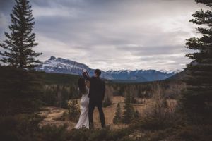 Bride and groom stand among pine trees as groom twirls bride with snowy mountains behind.