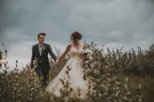 Bride and groom walk hand in hand through a wild meadow under dramatic overcast skies.