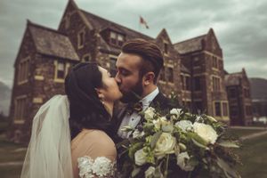 Newlyweds share a kiss in front of a historic stone building with a white floral bouquet.